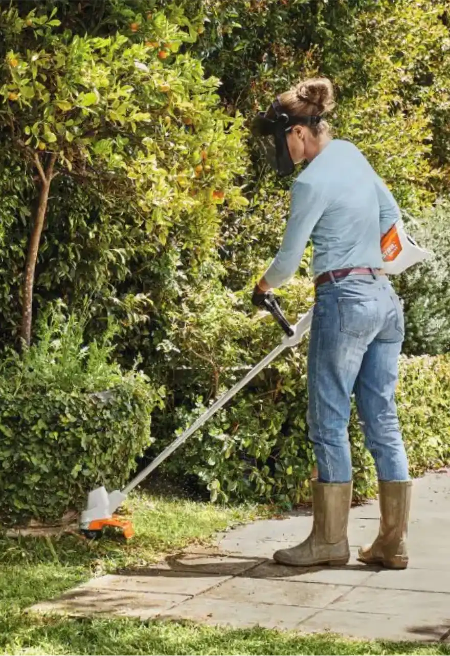 Jardineira usando roçadeira a bateria Stihl para aparar a grama ao lado de uma calçada