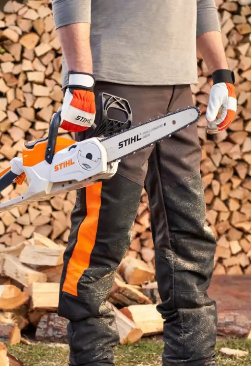 Operador com luvas e calça de proteção segurando motosserra Stihl diante de uma pilha de lenha